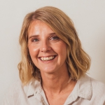 Emily, a white woman with short blonde hair wearing a beige shirt is posing for a photograph.