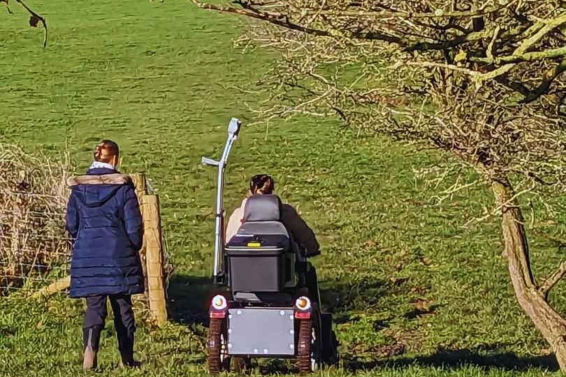 Two people are walking through a field, one standing the other using a mobility scooter, their backs are turned walking away from the camera. The field is green, a tree frames the picture.