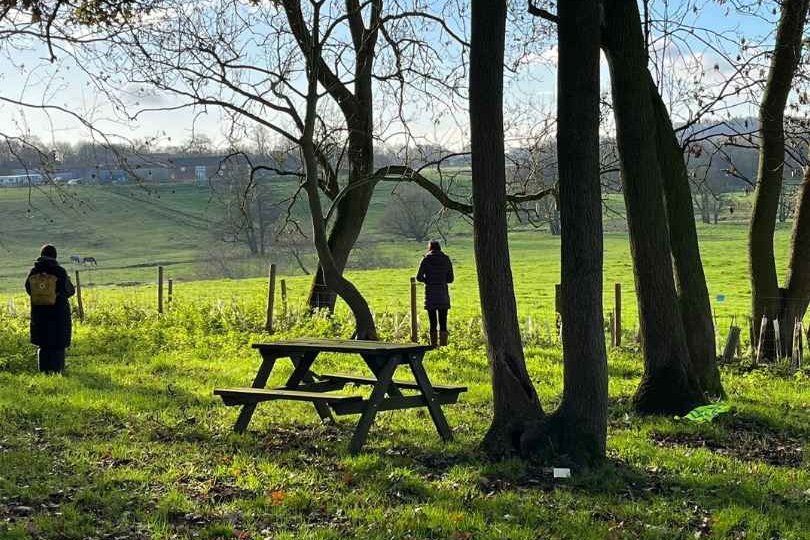 A woodland with beech trees and light coming through the canopy. A picnic table is in the centre of the image and two people stand with their backs turned looking into the distant rolling countryside.